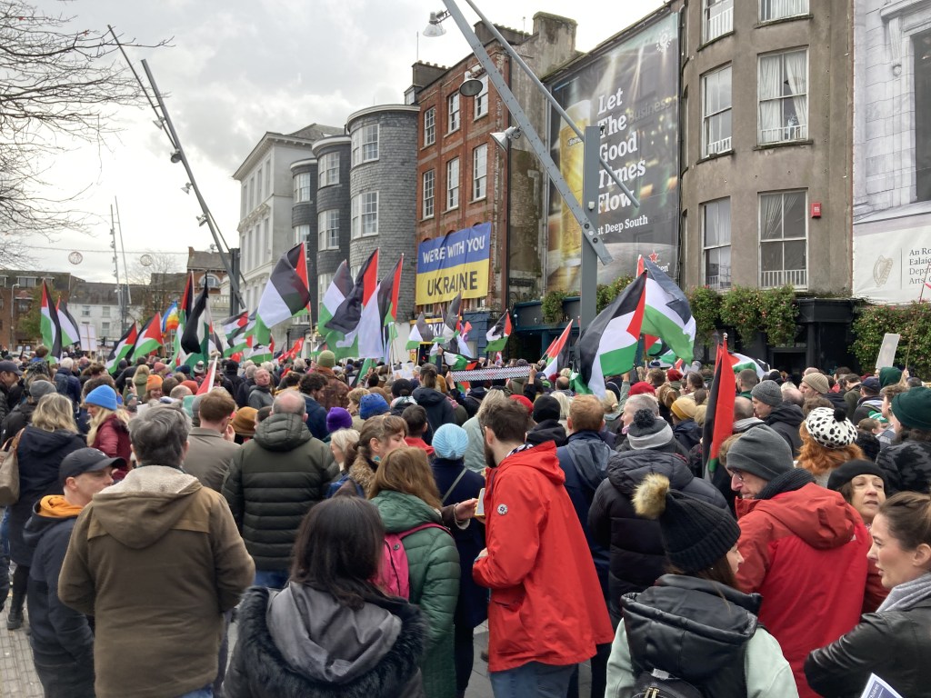 A crowd in Cork for a Free Palestine rally, with a large Ukrainian flag in the background with the words "We're with you Ukraine" printed on it.