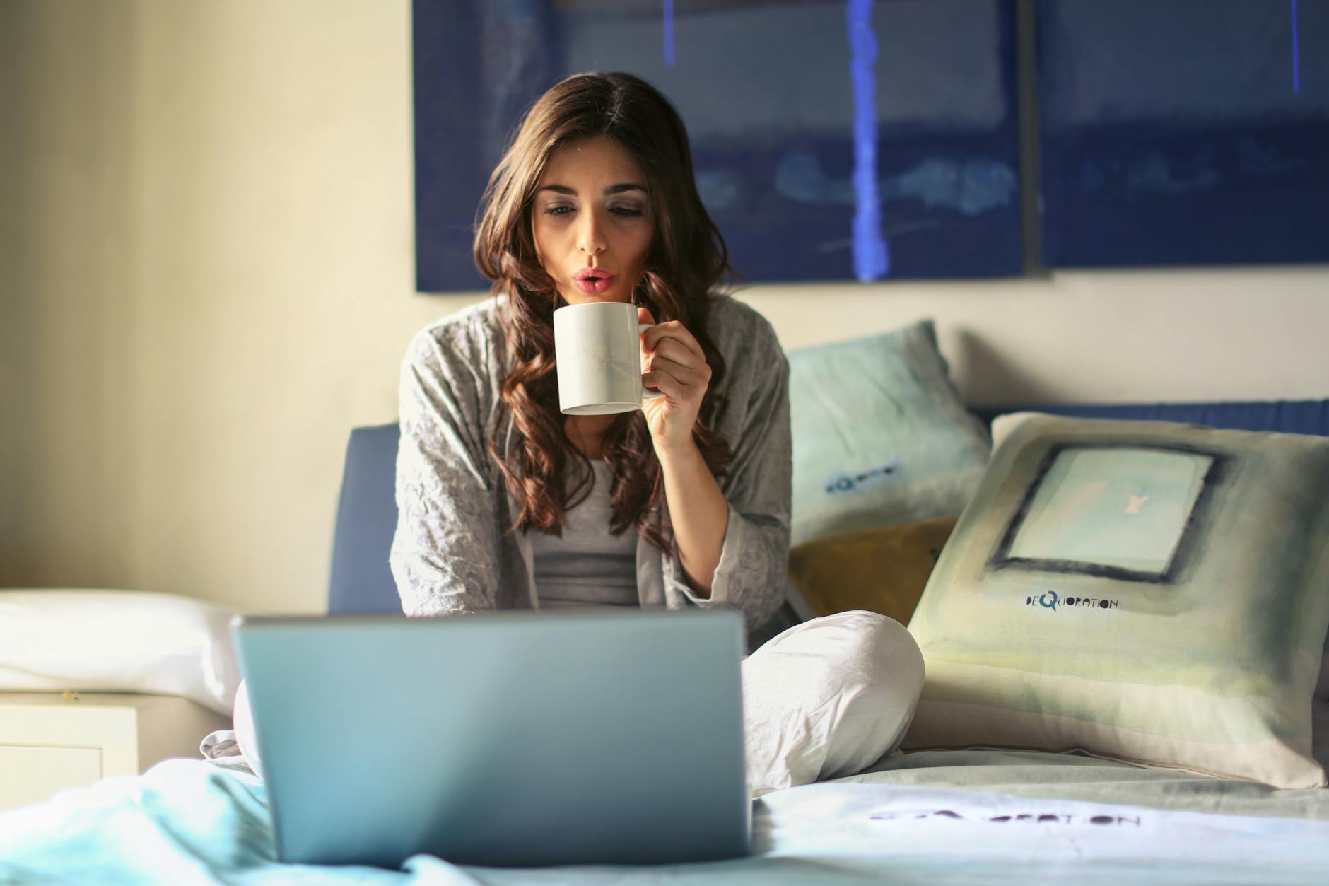 A woman sitting on a bed with a laptop and a cup of tea